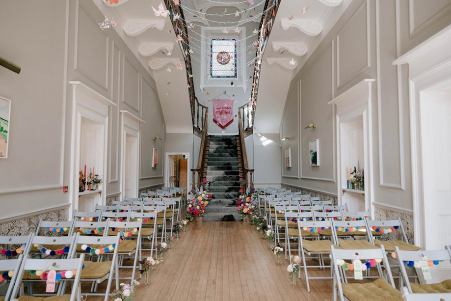 Wedding ceremony setup in the grand hall at Netherbyres House with aisle seating, staircase backdrop and rainbow pom-pom decor