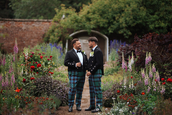 Two grooms in tartan trews walking through the walled garden at Netherbyres House during their Scottish wedding