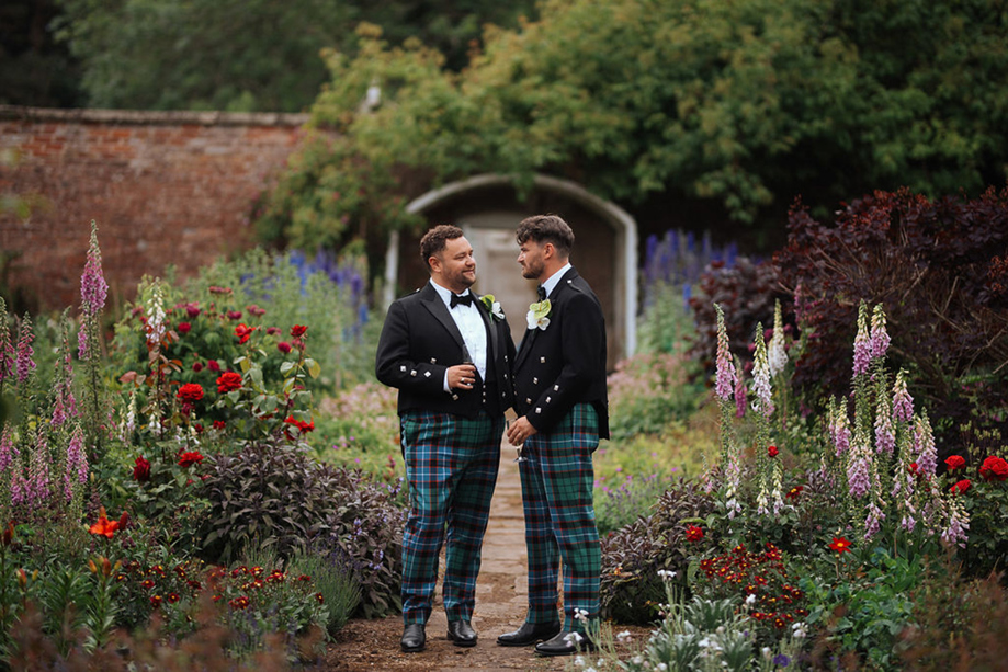 Two grooms in tartan trews walking through the walled garden at Netherbyres House during their Scottish wedding