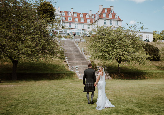 Bride looks back with sunglasses on walking with her groom towards Peebles Hydro Hotel