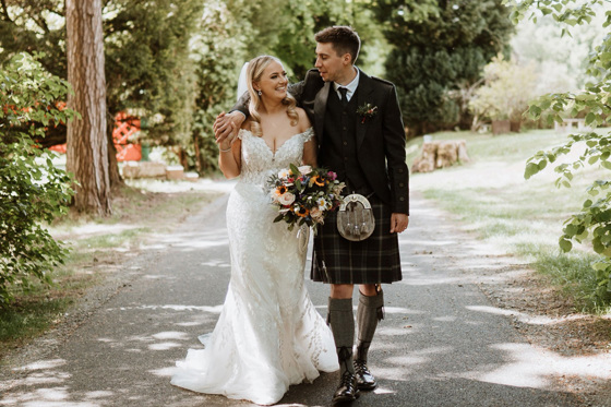 Bride and groom with arm round her shoulder smile at each other as they walk 