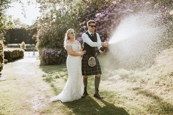 Bride and groom wearing sunglasses laugh as they spray champagne on Peebles Hydro Hotel grounds