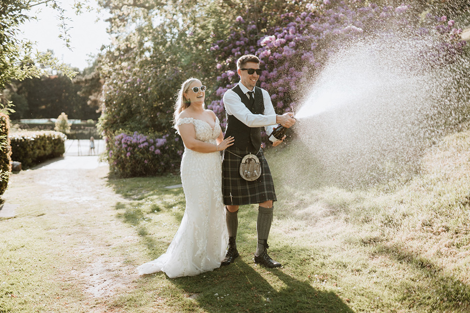 Bride and groom wearing sunglasses laugh as they spray champagne on Peebles Hydro Hotel grounds