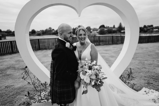 Bride and groom pose in front of white love heart in gardens of Riverside Lodge Hotel
