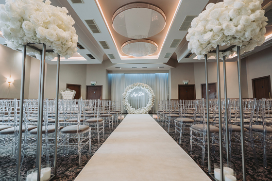Wedding ceremony set up with clear chairs, white flowers on silver stands and a white flower hoop at altar with "Aye Do" lights