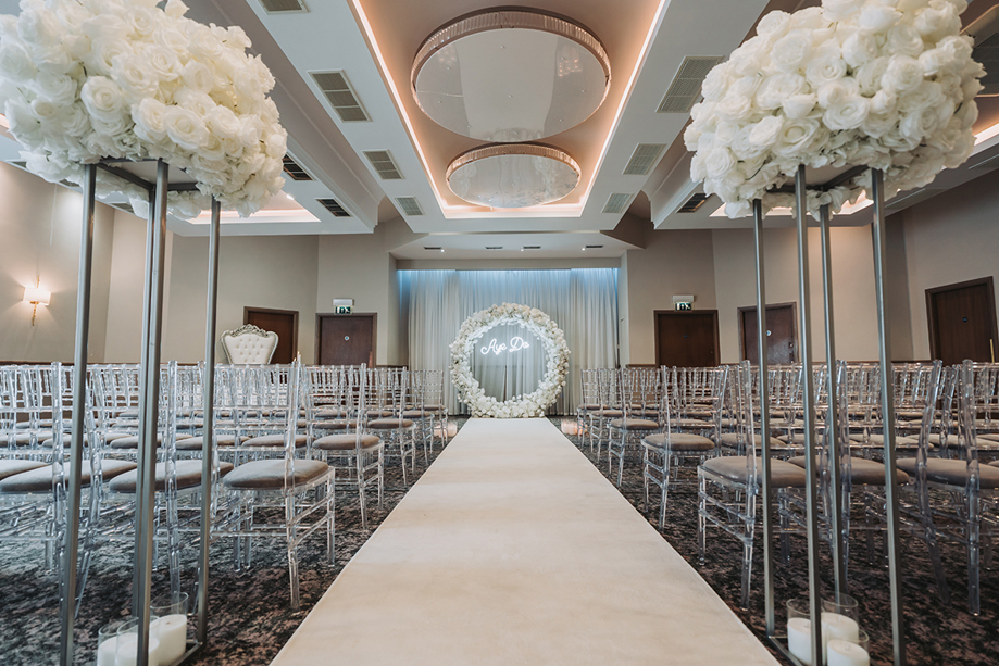 Wedding ceremony set up with clear chairs, white flowers on silver stands and a white flower hoop at altar with "Aye Do" lights