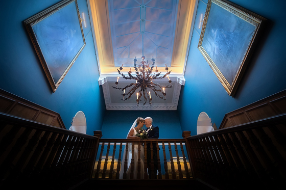 Bride and groom embrace at top of stairs at SCHLOSS Roxburghe with large chandelier and skylight above.