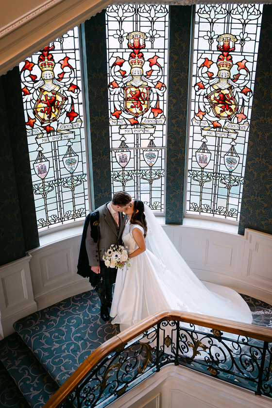 Bride and groom kissing on staircase landing at The Balmoral Hotel Edinburgh with stained glass windows behind