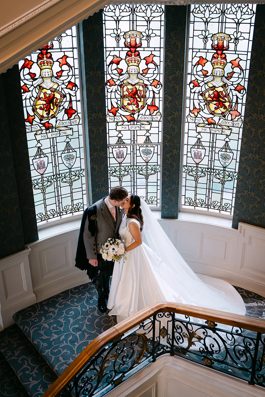 Bride and groom kissing on staircase landing at The Balmoral Hotel Edinburgh with stained glass windows behind