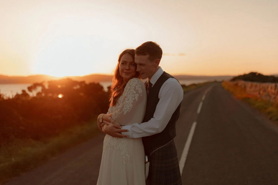 Bride and groom stand together on country road at sunset