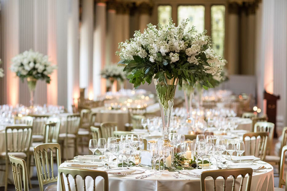View of wedding meal tables with bouquets of white flowers in tapered vases and gold chairs