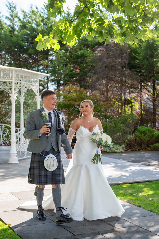 Bride and groom walk through gardens on sunny wedding day, groom is holding a pint of Guiness and the bride is holding her white rose bouquet