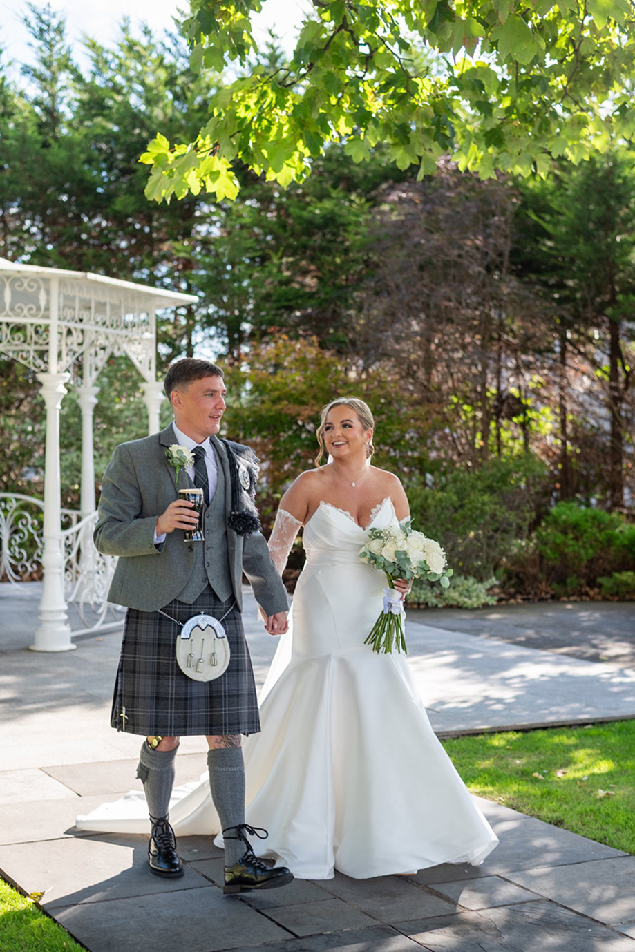 Bride and groom walk through gardens on sunny wedding day, groom is holding a pint of Guiness and the bride is holding her white rose bouquet
