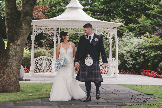 Bride and groom walk through gardens at Carlton Hotel in Prestwick, they are looking at each other and the bride is holding a baby blue bouquet