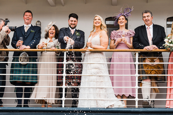 Guests smile as groom pops bottle of champagne aboard Fingal Hotel.