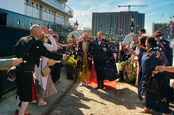 Two grooms laugh as they walk through confetti toss on the dock beside Fingal Hotel.