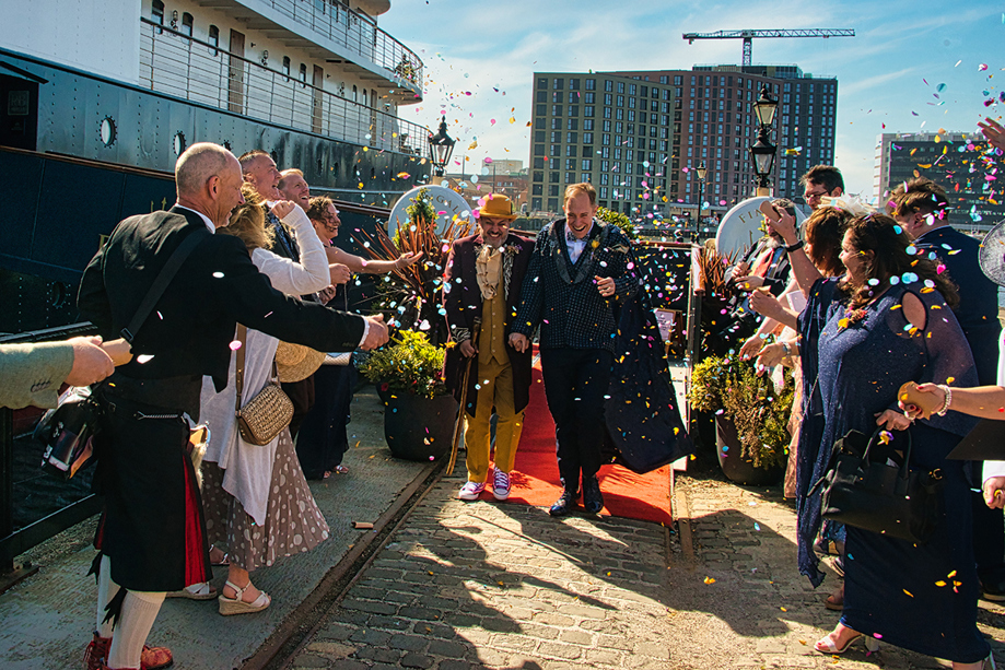Two grooms laugh as they walk through confetti toss on the dock beside Fingal Hotel.