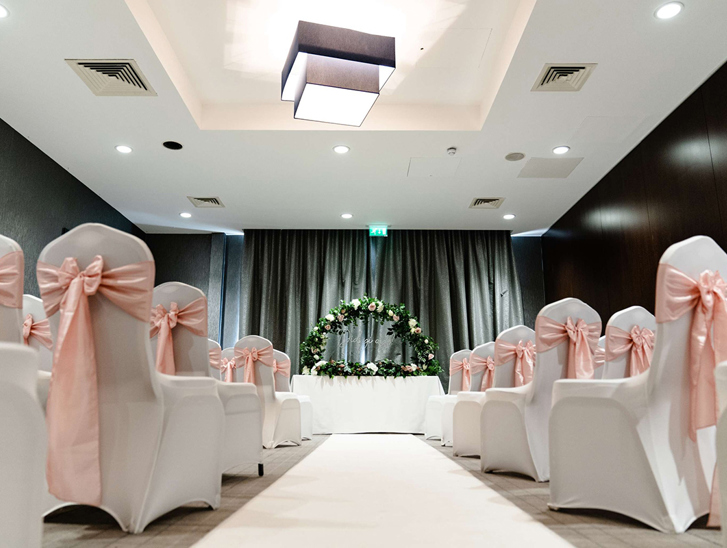Wedding ceremony room at Village Hotel Edinburgh with white aisle, pink chair bows and floral arch