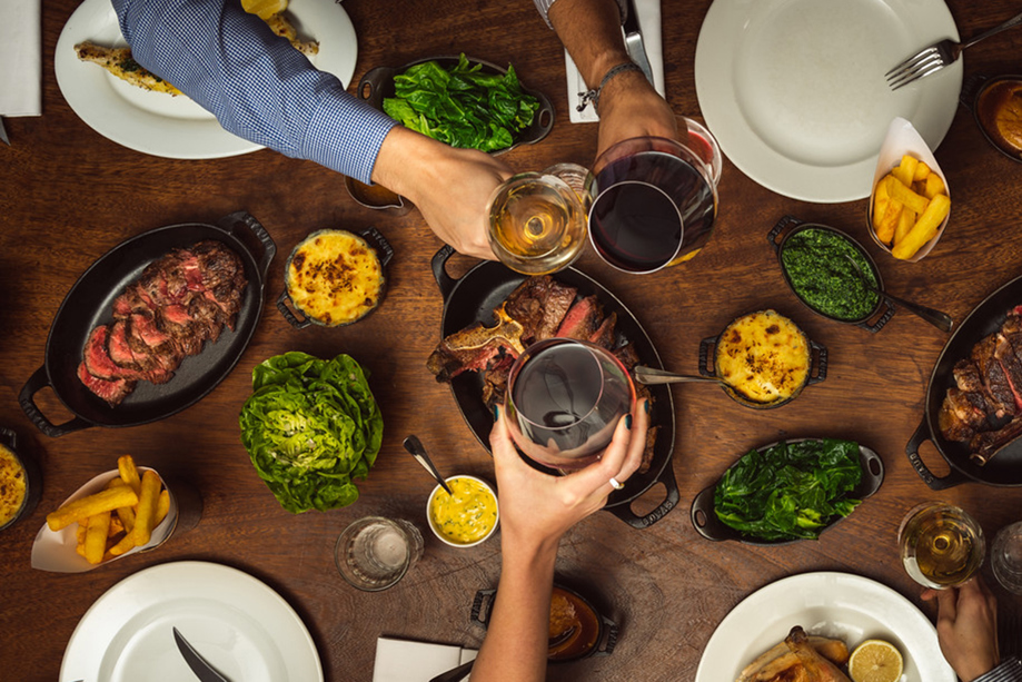 Top-down view of a Hawksmoor Edinburgh sharing meal with sliced steak, classic sides and guests toasting with wine.