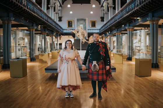 Bride and groom hold hands and face photographer in Kelvingrove Art Gallery following their University of Glasgow wedding.