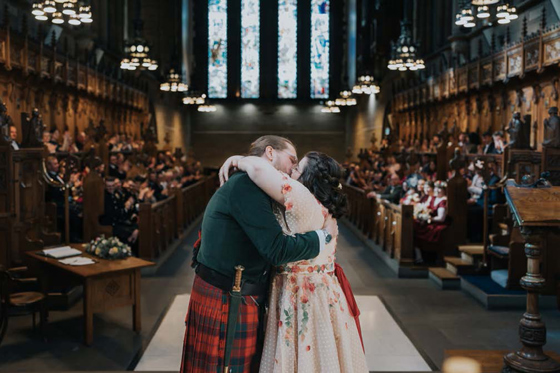 Bride wearing pink dress with flowers kisses new husband wearing a red and green kilt at the altar in the Memorial Chapel at the University of Glasgow.