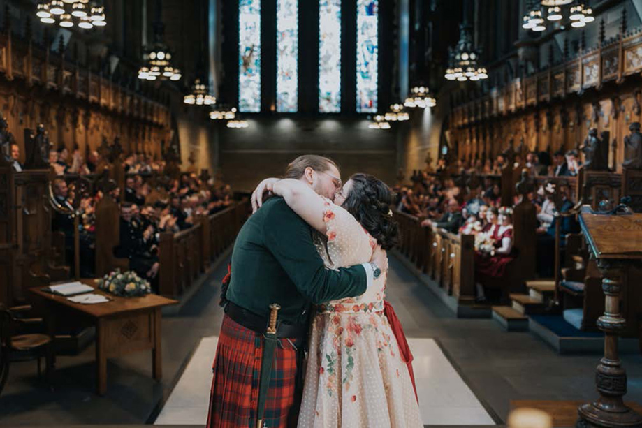 Bride wearing pink dress with flowers kisses new husband wearing a red and green kilt at the altar in the Memorial Chapel at the University of Glasgow.