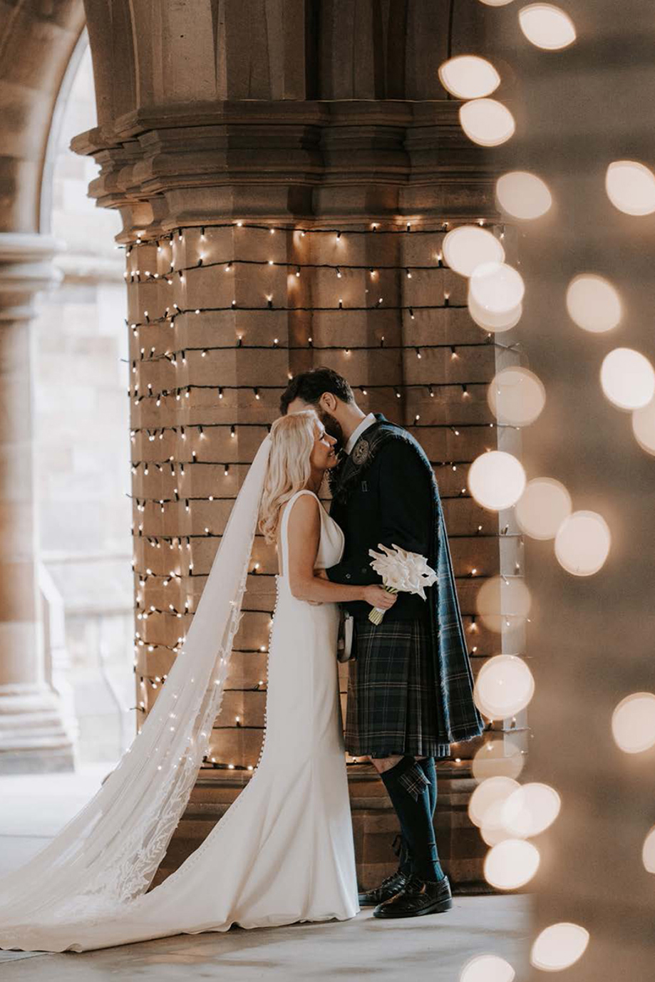 Bride and groom embrace in the fairy light-wrapped Cloisters at the University of Glasgow.