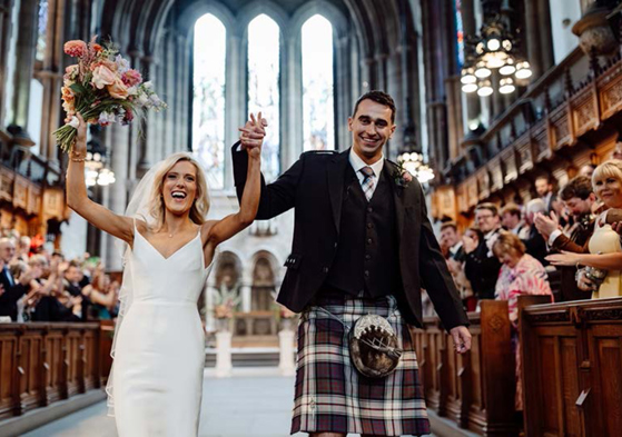 Bride and groom celebrate while walking back up the aisle hand-in-hand while guests clap in the Memorial Chapel at the University of Glasgow.