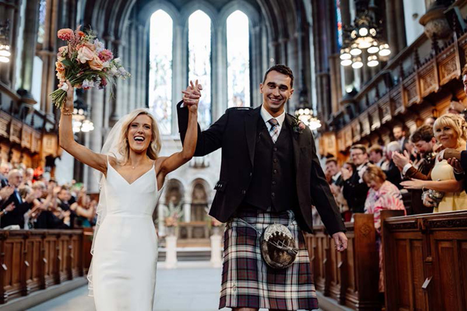 Bride and groom celebrate while walking back up the aisle hand-in-hand while guests clap in the Memorial Chapel at the University of Glasgow.