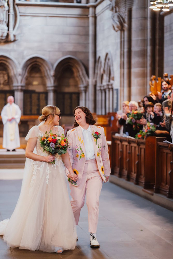 Two brides, one in a pink-tinted dress and one in a pink suit with flowers embroidered laugh together as they walk back up the aisle in the Memorial Chapel at the University of Glasgow.