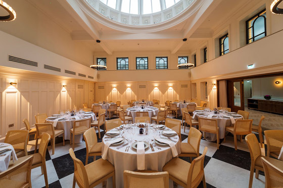 round tables with white linen and tableware set up for a wedding dinner below a glass dome on the ceiling