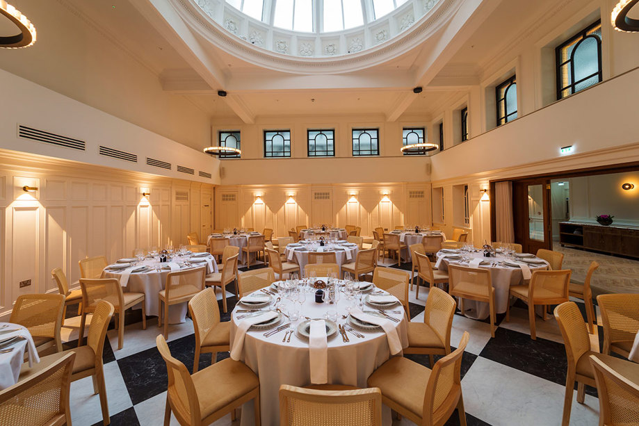 round tables with white linen and tableware set up for a wedding dinner below a glass dome on the ceiling