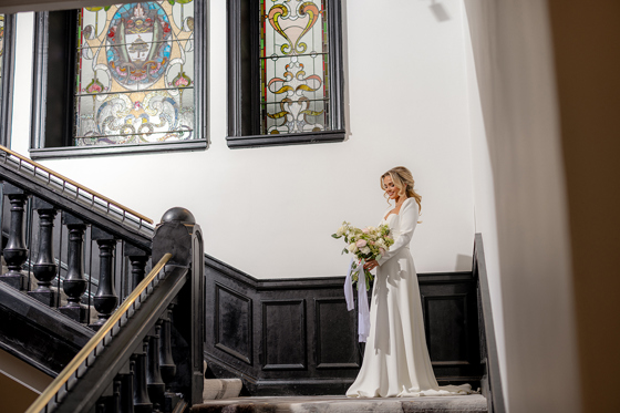 Bride holding bouquet stands on imposing The Collector's Hall staircase, with stained glass windows in background and dark panelling on wall.