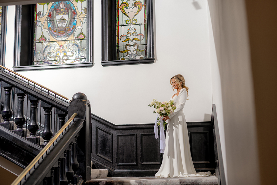 Bride holding bouquet stands on imposing The Collector's Hall staircase, with stained glass windows in background and dark panelling on wall.